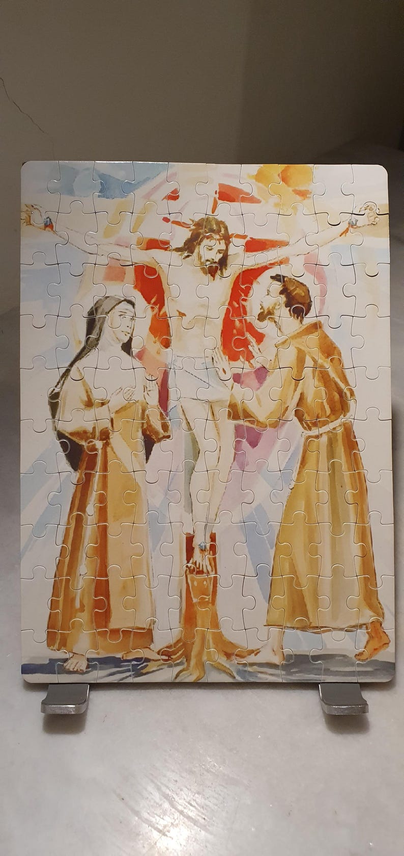 May include: A completed jigsaw puzzle depicting the crucifixion of Jesus Christ. The artwork features Jesus on the cross, with figures in robes and a vibrant sky background. The puzzle is displayed on a metal stand.
