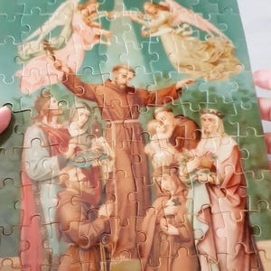 May include: A completed jigsaw puzzle featuring a religious scene with figures in brown robes and angels. The puzzle has a green background and a small box with the words "thank you" on top.