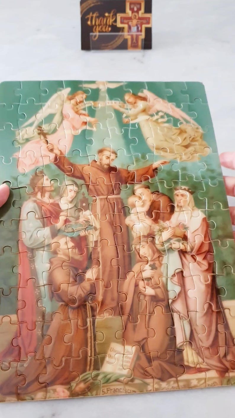 May include: A completed jigsaw puzzle featuring a religious scene with figures in brown robes and angels. The puzzle has a green background and a small box with the words "thank you" on top.