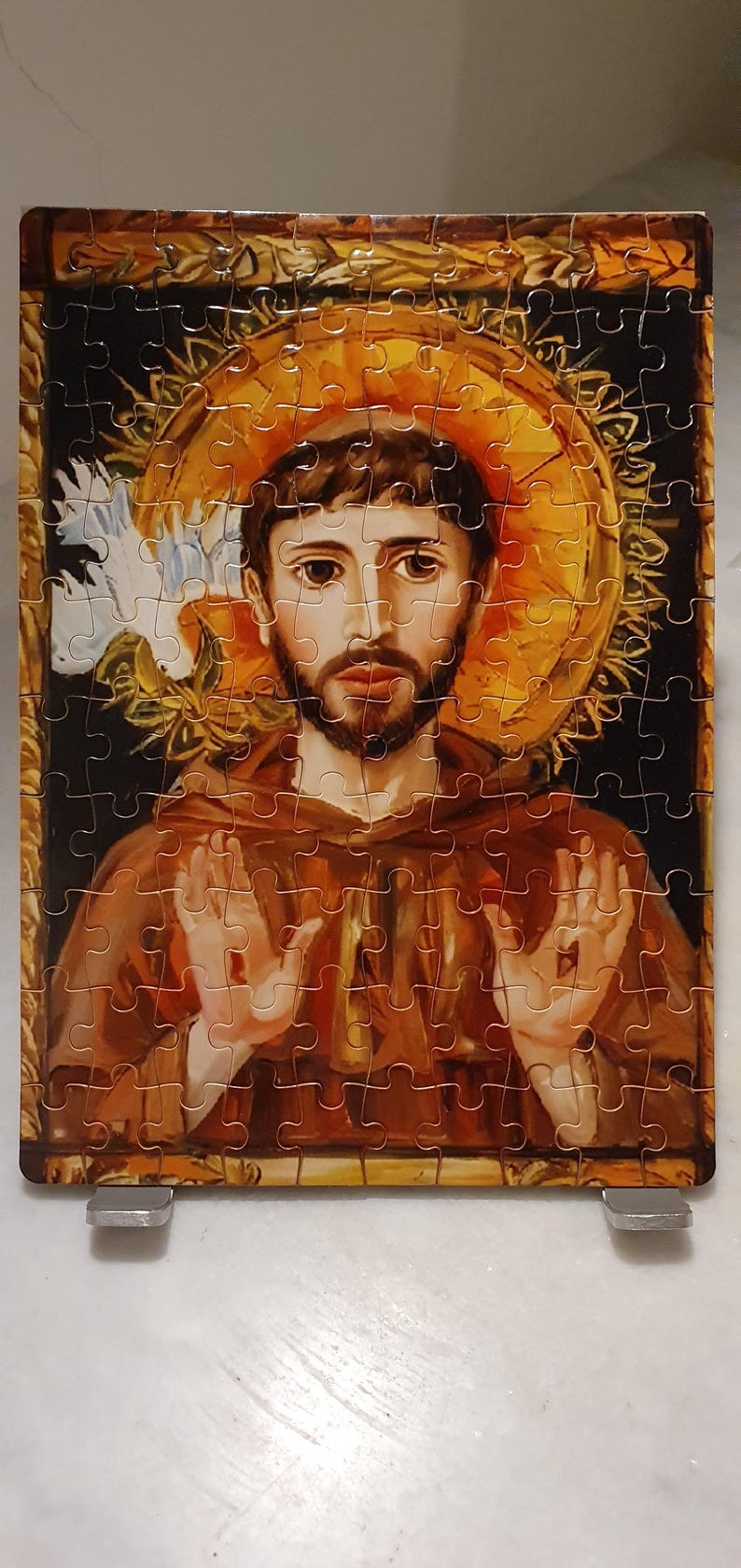 May include: A completed jigsaw puzzle featuring a religious figure with a halo. The puzzle depicts a man in brown robes with a beard, hands raised, and a golden halo. The puzzle is supported by two silver stands.