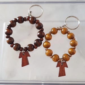 May include: Two wooden keyrings. One is dark brown, the other is light brown. Each keyring features a ring of wooden beads and a small, wooden cross pendant. The keyrings are displayed on a clear surface.