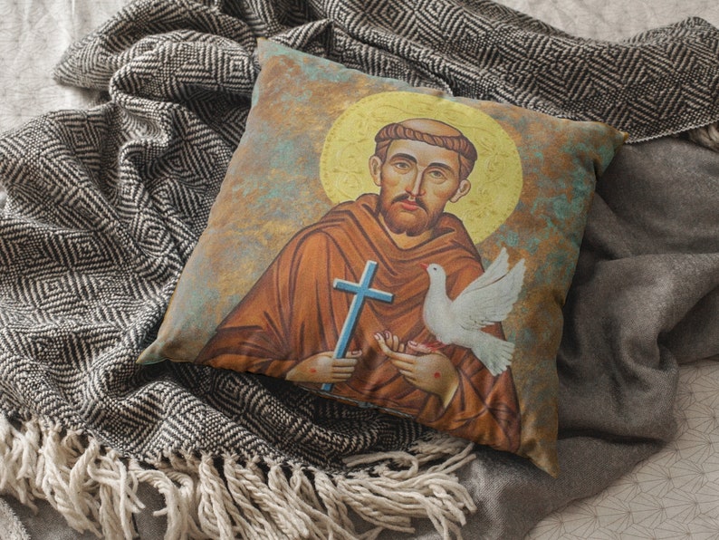 May include: A throw pillow with a vintage-style illustration of Saint Francis of Assisi. The image features Saint Francis holding a dove and a cross, with a halo behind his head. The background is a faded blue and green.