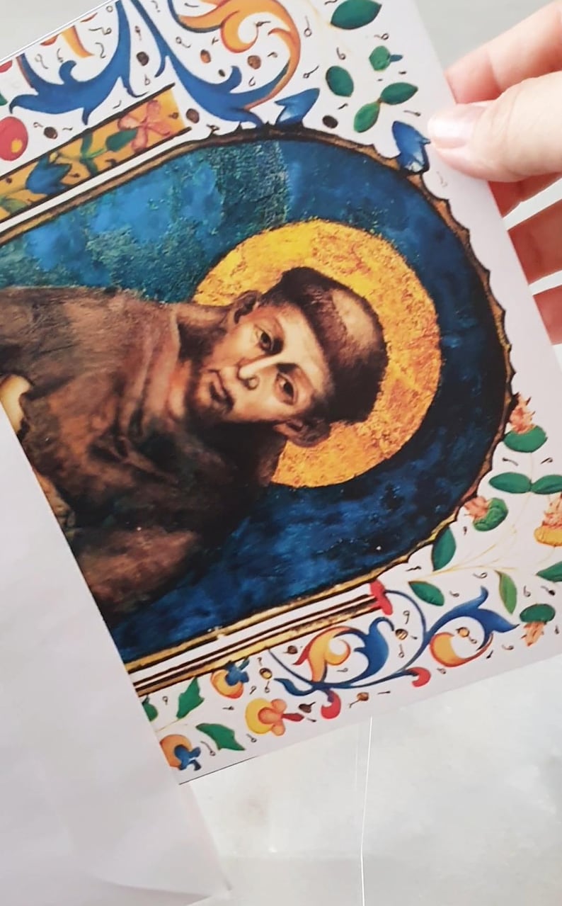 May include: A print of a religious figure with a golden halo, set against a blue background. The artwork features ornate floral and scrollwork borders in blue, yellow, and red. The figure is depicted in brown robes.