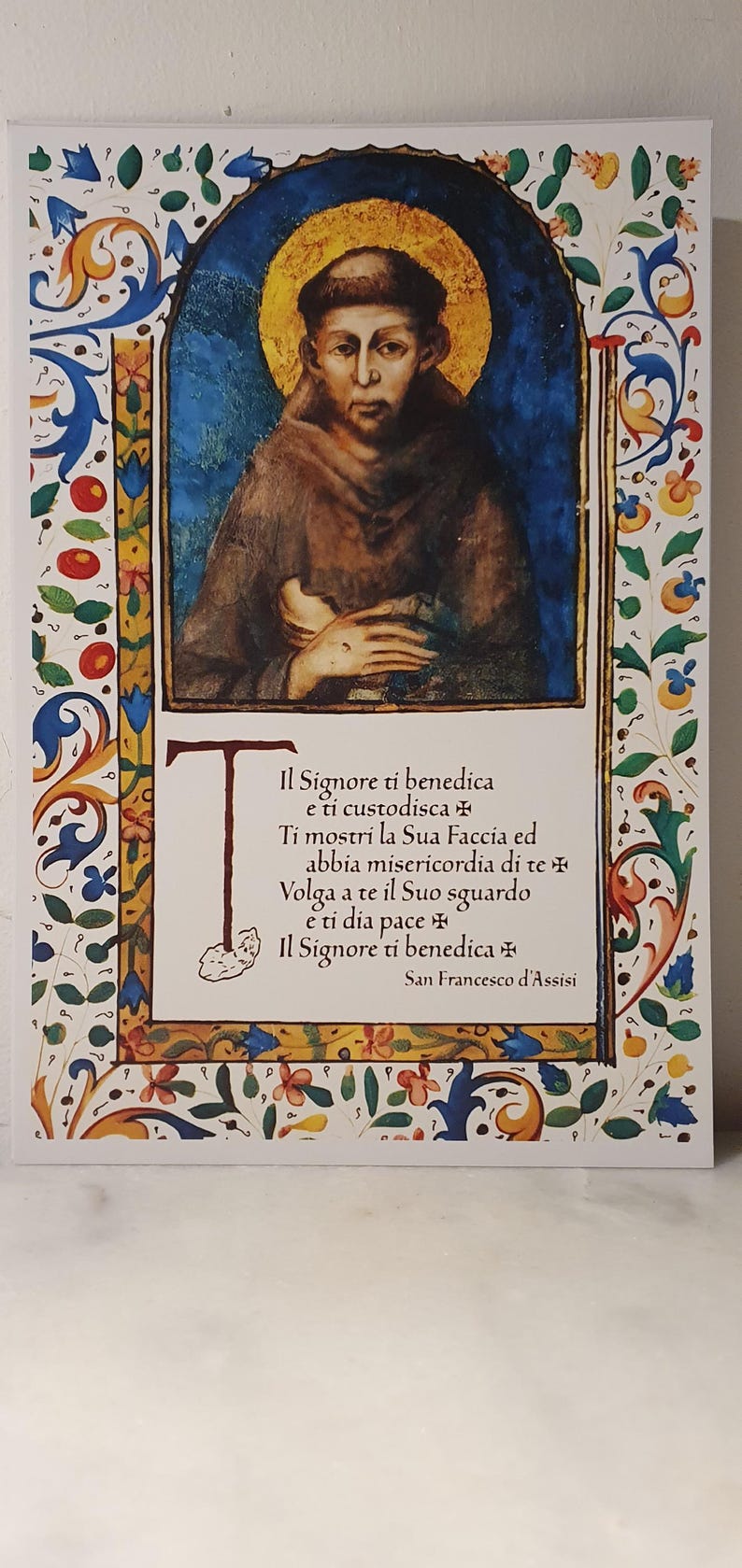 K&ouml;nnte beinhalten: Ein gerahmter Druck mit einer Illustration eines Mannes mit Heiligenschein, vor blauem Hintergrund. Der Druck enth&auml;lt italienischen Text und ist von einem dekorativen floralen Rand umgeben.