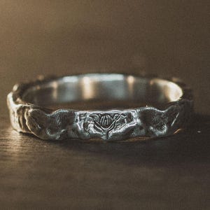 Può includere: Un anello d'argento con un design strutturato e organico. L'anello presenta un motivo floreale dettagliato e una finitura martellata. L'anello è esposto su una superficie di legno scuro, evidenziando i suoi dettagli e la sua maestria. Un gioiello.