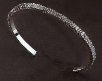May include: A silver-colored cuff bracelet with an open design. The outer surface features a textured, vertical ridged pattern that contrasts with the smooth, polished interior finish of the metal band, creating a distinct visual appearance.