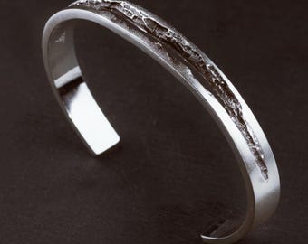 May include: A silver-colored cuff bracelet with a smooth, polished finish on the outer edges and a central, recessed channel featuring a dark, rugged, and uneven textured surface that runs along the entire length of the open-ended metal band.