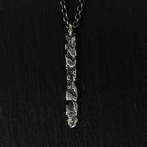 May include: A silver-colored pendant necklace with a textured, elongated bar design. The pendant hangs from a delicate chain. The necklace is displayed against a dark background.