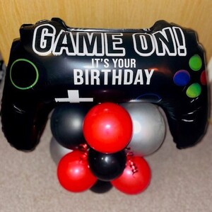Könnte beinhalten: Ein schwarz-weißer Ballon in Form eines Videospiel-Controllers mit dem Text "GAME ON! It's your birthday". Der Ballon ist von kleineren roten, schwarzen und silbernen Ballons umgeben.