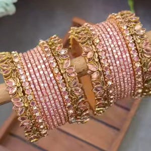May include: Two sets of pink and gold bangles. Each set has multiple bangles with a pink, gem-like center and gold floral designs on the edges. The bangles are displayed on a wooden dowel.