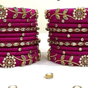 Op de afbeelding: Een set van 12 roze zijden draad armbanden met gouden en witte versieringen. De armbanden zijn versierd met kleine bladeren, strass steentjes en parels.