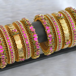 Puede incluir: Brazaletes dorados con diseños intrincados. Cada brazalete presenta una fila de gemas rosas vibrantes, pequeños cristales blancos y detalles de monedas doradas. Los brazaletes se exhiben en un soporte negro sobre un fondo blanco suave.