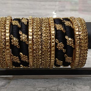 Op de afbeelding: Een verzameling goudkleurige armbanden met ingewikkelde ontwerpen, afgewisseld met zwarte armbanden versierd met bloempatronen. De armbanden worden gepresenteerd op een zwarte standaard, die hun sierlijke details en contrasterende kleuren benadrukt.