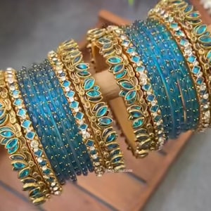 May include: A collection of teal and gold bangles. The bangles are arranged on a wooden dowel. The teal bangles are accented with gold filigree and small, clear gemstones. The bangles have a delicate, ornate design.
