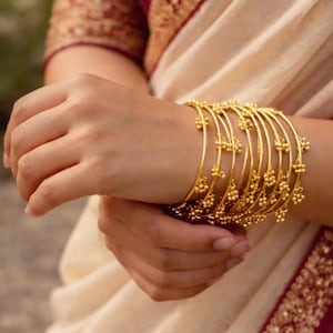 May include: A set of gold bangles, each featuring small, decorative clusters. The bangles are stacked on a wrist, highlighting their detailed design. The background includes cream-coloured fabric with burgundy and gold accents.