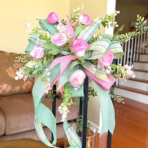 May include: A black metal lantern with a glass cylinder holding a green candle. The lantern is decorated with pink roses, green leaves, and ribbons in pink and green plaid. The lantern is on a glass table.