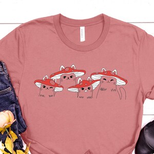May include: A heather mauve t-shirt with a graphic of four cats wearing red mushroom hats. The cats have different expressions and the text "meow meow" is written below them.