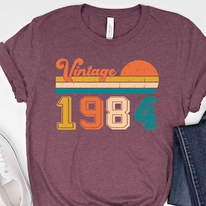 Peut inclure: Un t-shirt marron avec un design vintage 1984. Le design pr&eacute;sente un coucher de soleil avec un fond ray&eacute; et l'ann&eacute;e 1984 dans une police r&eacute;tro color&eacute;e.