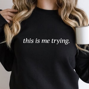 May include: Black sweatshirt with the text "this is me trying." in white lettering.