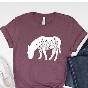 May include: A maroon t-shirt with a white silhouette of a donkey with flowers inside the outline. The shirt is laid out on a white surface with a pair of white sneakers and a pair of blue jeans.