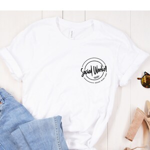 Puede incluir: Camiseta blanca con un gr&aacute;fico circular negro que dice "Social Worker" sobre un fondo de madera blanca con jeans azules, flores rosas y sandalias marrones.