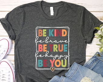 May include: A dark gray t-shirt with a white graphic that says "Be Kind, Be Brave, Be True, Be Happy, Be You" in colorful letters. The graphic is surrounded by a white frame with a colorful border. A small heart with a leopard print design is in the center of the frame.