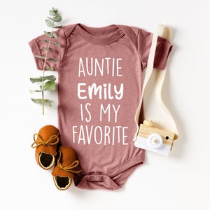 May include: A mauve onesie with the text "Auntie Emily is my favorite" in white. The onesie is lying on a white surface with a wooden toy camera and brown leather baby shoes.