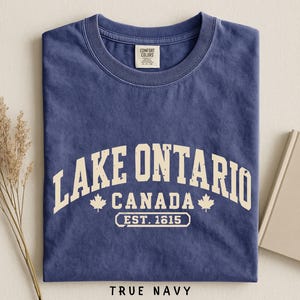 May include: A true navy blue short-sleeve t-shirt with a crew neckline. The front features cream-colored distressed text that reads LAKE ONTARIO CANADA EST. 1615, flanked by two small maple leaf icons. The shirt has a Comfort Colors brand label.