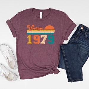 Może przedstawiać: Bordowy t-shirt z vintage'owym nadrukiem z 1979 roku przedstawiającym zach&oacute;d słońca i pasy w kolorze pomarańczowym, ż&oacute;łtym, turkusowym i zielonym.