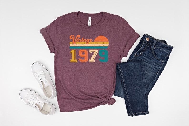 Może przedstawiać: Bordowy t-shirt z vintage'owym nadrukiem z 1979 roku przedstawiającym zach&oacute;d słońca i pasy w kolorze pomarańczowym, ż&oacute;łtym, turkusowym i zielonym.