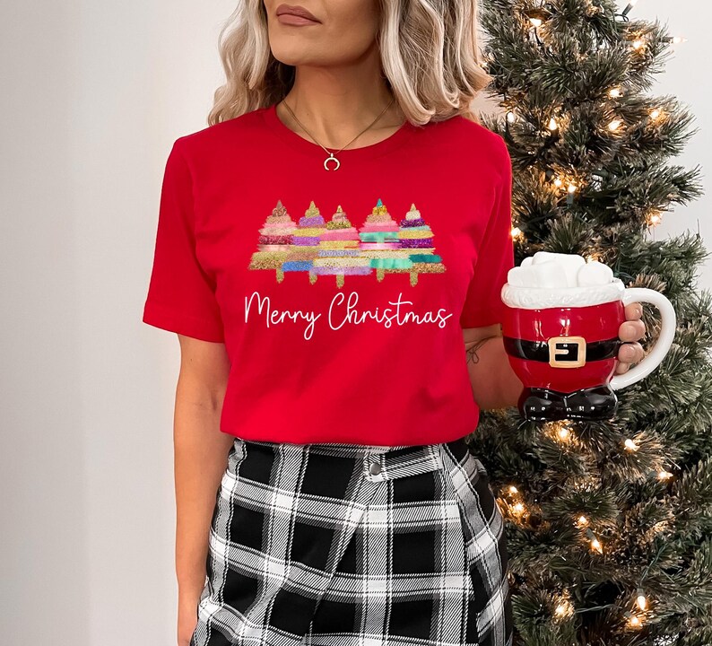 K&ouml;nnte beinhalten: Rotes T-Shirt mit einem Motiv von bunten Weihnachtsb&auml;umen und dem Text "Merry Christmas".
