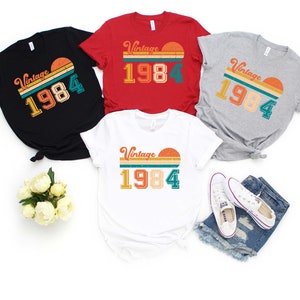 Peut inclure: Quatre t-shirts avec un design vintage 1984. Les t-shirts sont en noir, rouge, gris et blanc. Chaque t-shirt pr&eacute;sente un graphique de coucher de soleil avec le texte "Vintage 1984" dans une police r&eacute;tro.
