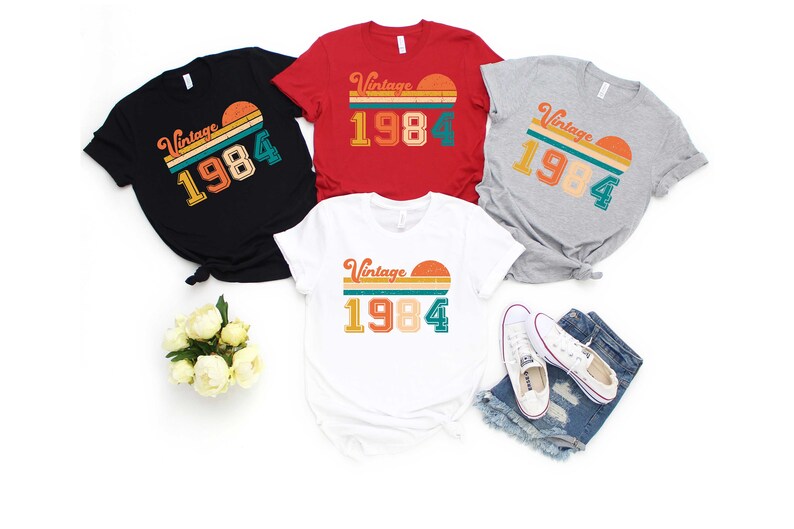Peut inclure: Quatre t-shirts avec un design vintage 1984. Les t-shirts sont en noir, rouge, gris et blanc. Chaque t-shirt pr&eacute;sente un graphique de coucher de soleil avec le texte "Vintage 1984" dans une police r&eacute;tro.