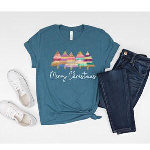 K&ouml;nnte beinhalten: T-Shirt in Tealblau mit einem Motiv von drei bunten Weihnachtsb&auml;umen und dem Text "Frohe Weihnachten".