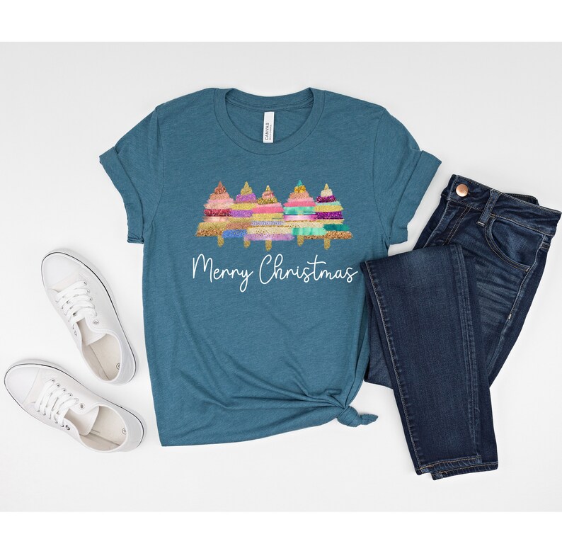 K&ouml;nnte beinhalten: T-Shirt in Tealblau mit einem Motiv von drei bunten Weihnachtsb&auml;umen und dem Text "Frohe Weihnachten".