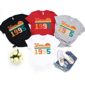 May include: A collection of t-shirts in black, red, white, and gray, each featuring a retro design with the text "Vintage 1995" and a sunset graphic. The shirts are casual wear, suitable for everyday use.