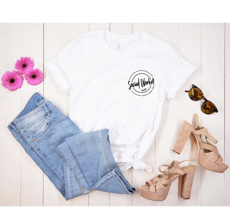 Puede incluir: Camiseta blanca con un gr&aacute;fico circular negro que dice "Social Worker" sobre un fondo de madera blanca con jeans azules, flores rosas y sandalias marrones.