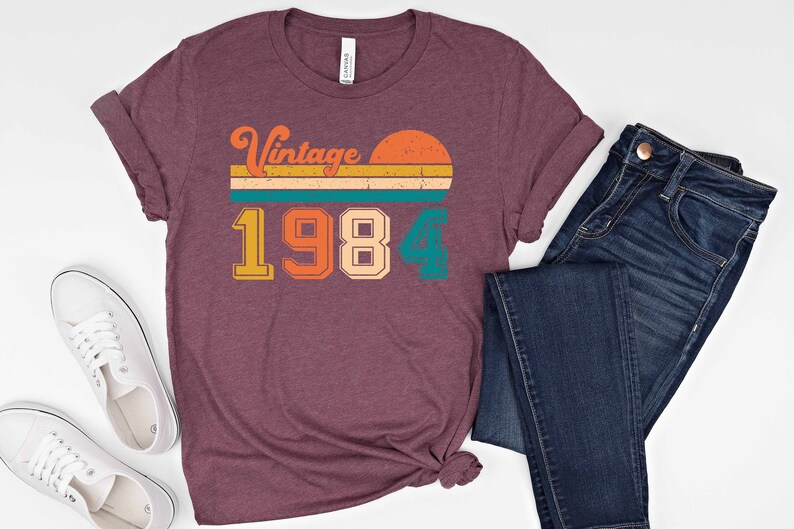 Peut inclure: Un t-shirt marron avec un design vintage 1984. Le design pr&eacute;sente un coucher de soleil avec un fond ray&eacute; et l'ann&eacute;e 1984 dans une police r&eacute;tro color&eacute;e.