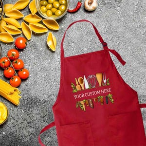May include: Red kitchen apron with a pocket and a design of kitchen utensils and the text "YOUR CUSTOM HERE" printed on the front.