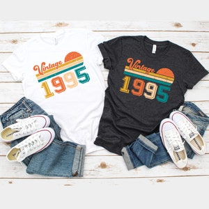 May include: Two t-shirts, one white and one dark gray, with the text "Vintage 1995" in a retro design. The shirts are paired with blue jeans and white sneakers. The design features a sunset graphic.