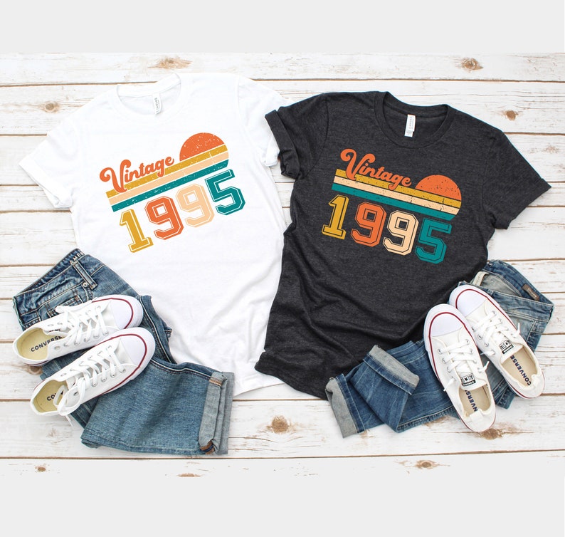 May include: Two t-shirts, one white and one dark gray, with the text "Vintage 1995" in a retro design. The shirts are paired with blue jeans and white sneakers. The design features a sunset graphic.
