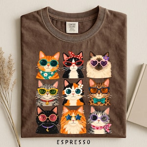 A t-shirt with a graphic design of nine cats wearing sunglasses and accessories, arranged in a 3x3 grid.