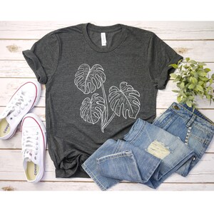May include: A dark gray t-shirt with a white outline of three monstera leaves. The shirt is laid out on a white wooden surface with a pair of white sneakers and blue jeans.
