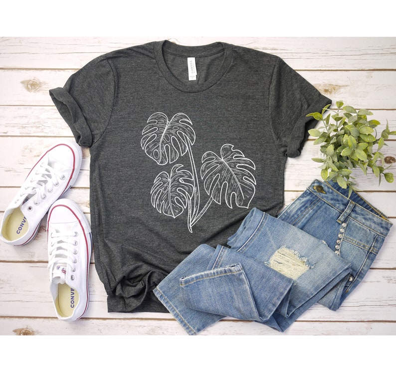 May include: A dark gray t-shirt with a white outline of three monstera leaves. The shirt is laid out on a white wooden surface with a pair of white sneakers and blue jeans.