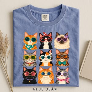A blue t-shirt with a collage of nine cat illustrations wearing sunglasses and accessories, arranged in a 3x3 grid.