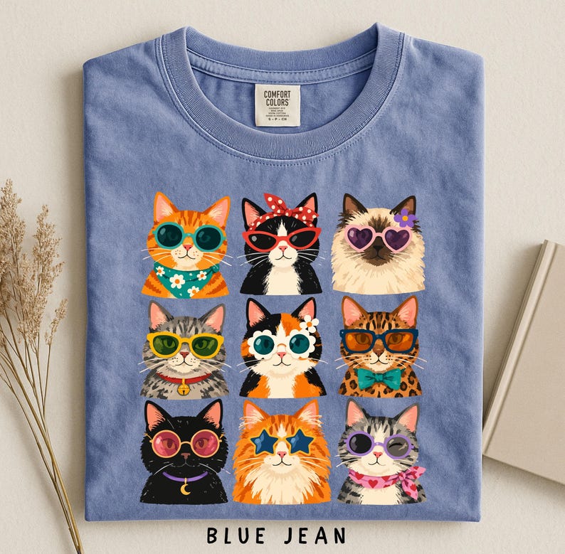 A blue t-shirt with a collage of nine cat illustrations wearing sunglasses and accessories, arranged in a 3x3 grid.