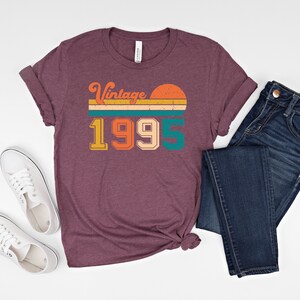 May include: A maroon t-shirt with the text "Vintage 1995" in a retro design, paired with blue jeans and white sneakers. The shirt features a sunset graphic and is likely a vintage-themed apparel item.