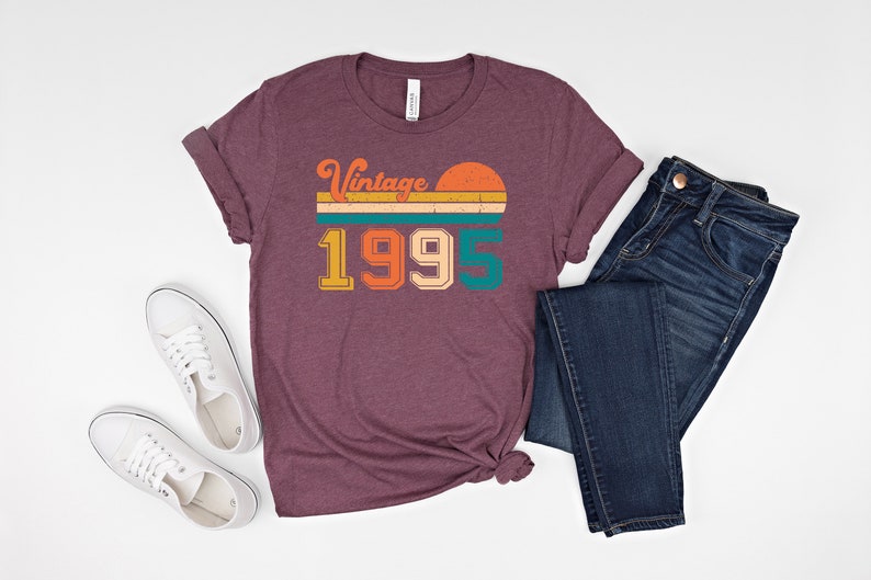 May include: A maroon t-shirt with the text "Vintage 1995" in a retro design, paired with blue jeans and white sneakers. The shirt features a sunset graphic and is likely a vintage-themed apparel item.