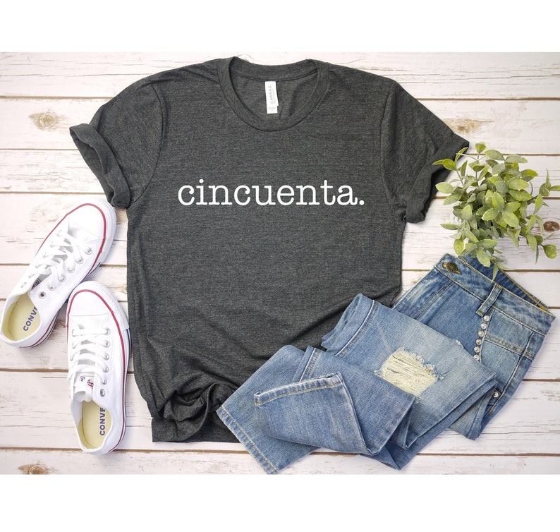 May include: A dark grey t-shirt with the word "cincuenta" printed in white on the front. The shirt is laid flat on a white wooden surface with a pair of white trainers and a pair of blue jeans.