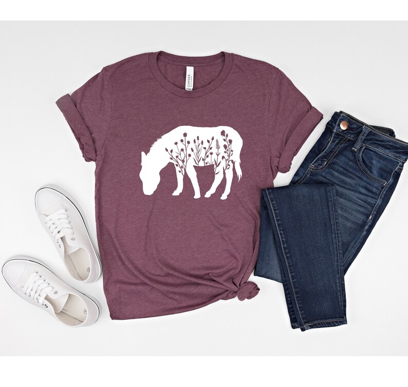 May include: A maroon t-shirt with a white silhouette of a donkey with flowers inside the outline. The shirt is laid out on a white surface with a pair of white sneakers and a pair of blue jeans.